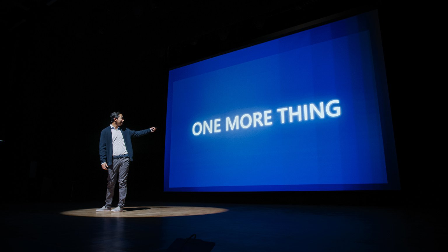 Live Event with Brand New Products Reveal: Keynote Speaker Presents New Device to Audience. Movie Theater Screen Shows Text -One More Thing- and Top Highlights.